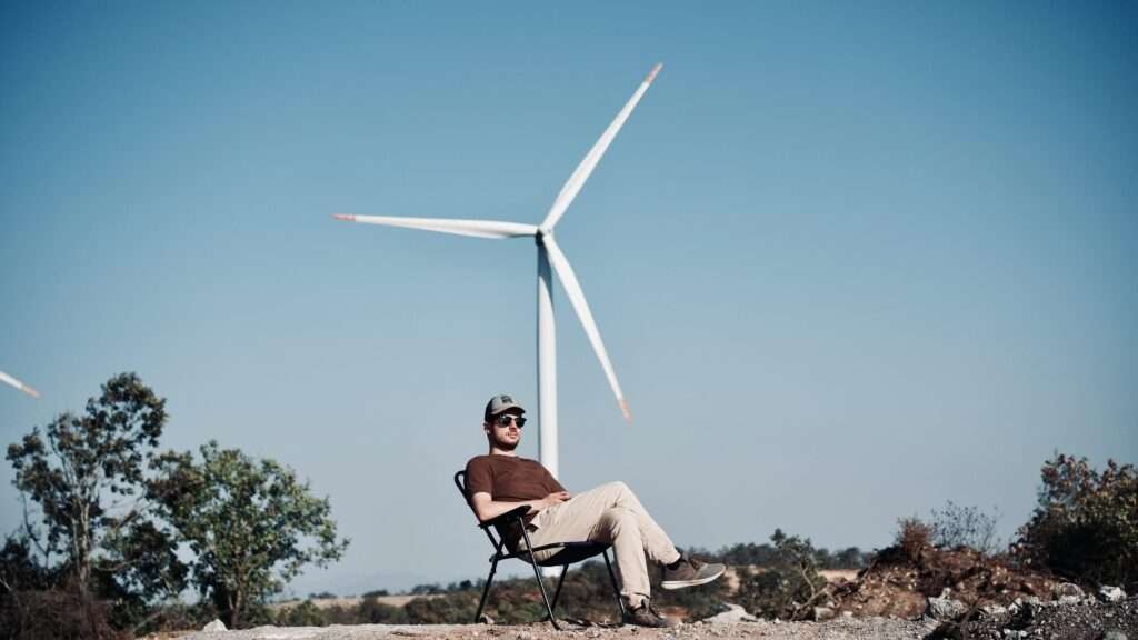 Man sitting outdoors on a folding chair near a large wind turbine under a clear blue sky